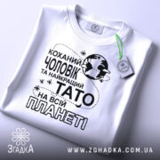 Футболка чоловік тато білого кольору, зображена зверху, на столі, фон світлий, принт чорного кольору з текстом "Коханий чоловік та найкращий Тато на всій планеті" та етикетка з зеленим шнурком.