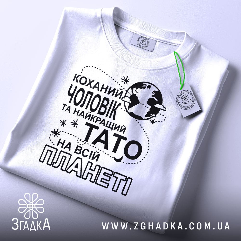 Футболка чоловік тато білого кольору, зображена зверху, на столі, фон світлий, принт чорного кольору з текстом "Коханий чоловік та найкращий Тато на всій планеті" та етикетка з зеленим шнурком.
