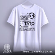 Футболка чоловік тато білого кольору, зображена з фронтального ракурсу, підвішена на вішалці, фон світлий, принт чорного кольору з текстом "Коханий чоловік та найкращий Тато на всій планеті".