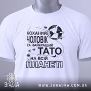 Футболка чоловік тато білого кольору, зображена зверху, на столі, фон світлий, принт чорного кольору з текстом "Коханий чоловік та найкращий Тато на всій планеті" та етикетка з зеленим шнурком.
