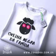 Футболка чорна вівця, біла, зображена зверху на столі, фон світло-сірий, принт з чорним овечим обличчям у рожевих окулярах та написом 'ovelha negra da família', з етикеткою, що звисає.