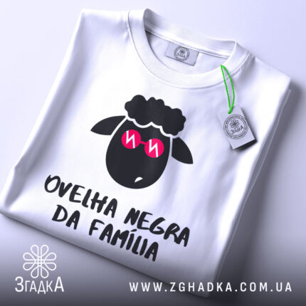 Футболка чорна вівця, біла, зображена зверху на столі, фон світло-сірий, принт з чорним овечим обличчям у рожевих окулярах та написом 'ovelha negra da família', з етикеткою, що звисає.