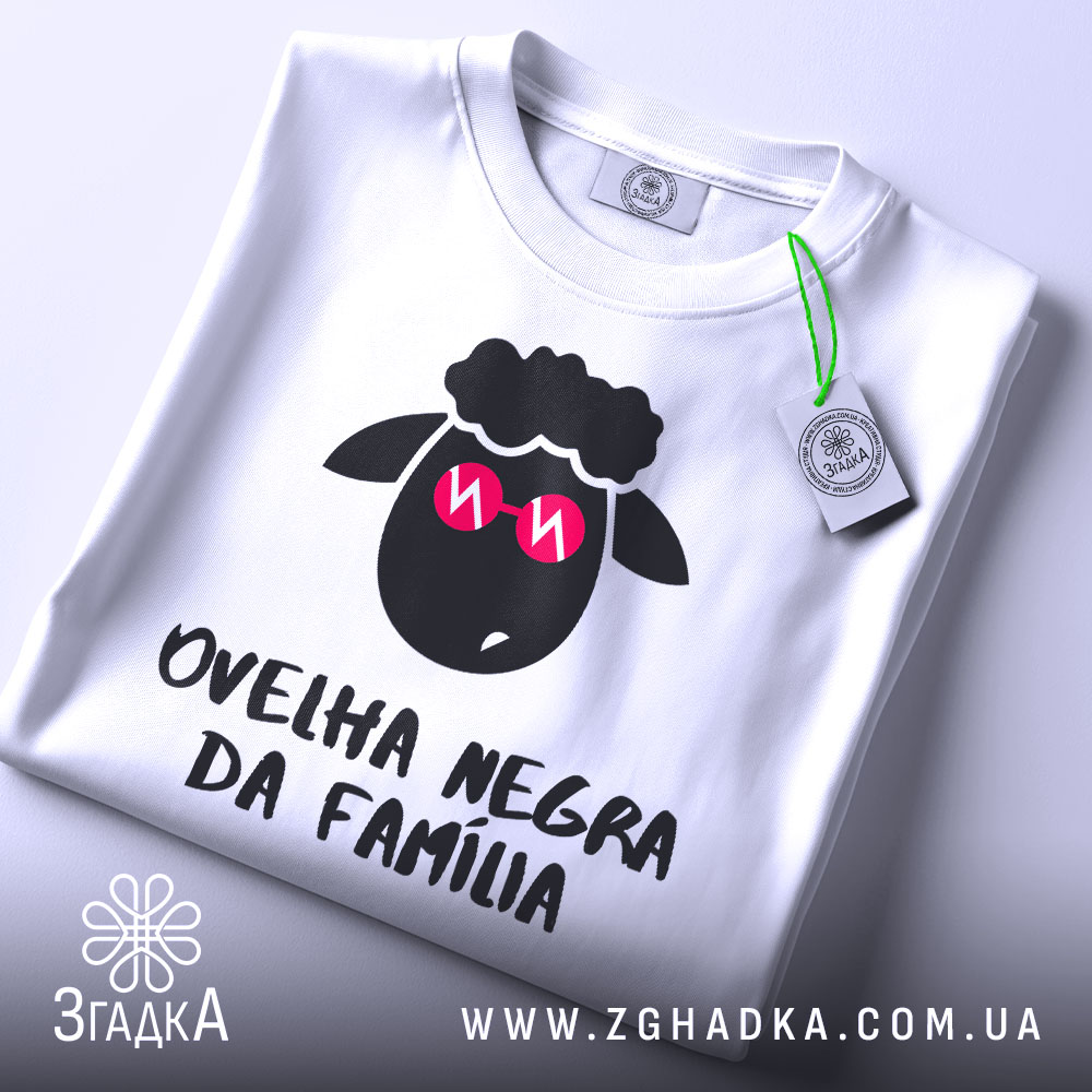 Футболка чорна вівця, біла, зображена зверху на столі, фон світло-сірий, принт з чорним овечим обличчям у рожевих окулярах та написом 'ovelha negra da família', з етикеткою, що звисає.