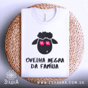 Футболка чорна вівця, біла, зображена зверху на плетеному кошику, фон світлий, принт з чорним овечим обличчям у рожевих окулярах та написом 'ovelha negra da família'.
