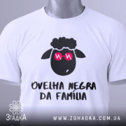 Футболка чорна вівця, біла, зображена зверху на столі, фон світло-сірий, принт з чорним овечим обличчям у рожевих окулярах та написом 'ovelha negra da família', з етикеткою, що звисає.