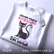 Футболка чорній кішці всеодно, біла, зображена з верхнього ракурсу, фон світлий, принт з чорним котом, рожевим текстом та біркою з зеленим шнурком.