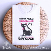 Футболка чорній кішці всеодно, біла, зображена зверху на плетеній кошику, фон нейтральний, принт з чорним котом, текстом та видимим ярликом.