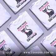 Футболка чорній кішці всеодно, біла, зображена з фронтального ракурсу на манекені, фон нейтральний, принт з чорним котом з рожевим бантиком та текстом.