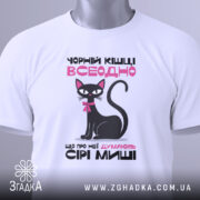 Футболка чорній кішці всеодно, біла, зображена з верхнього ракурсу, фон світлий, принт з чорним котом, рожевим текстом та біркою з зеленим шнурком.