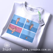 Футболка чорний гумор, біла футболка на столі, зображення принта з чотирма сценами, вид зверху, деталі рук, що з'являються з води, одна рука тягнеться до іншої, яка піднімається.