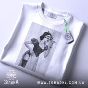 Футболка чорний гумор, біла, зображена зверху, на фоні світло-сірого кольору, принт з жінкою в костюмі, з написом 'BITCH', на футболці є етикетка з зеленим шнурком.