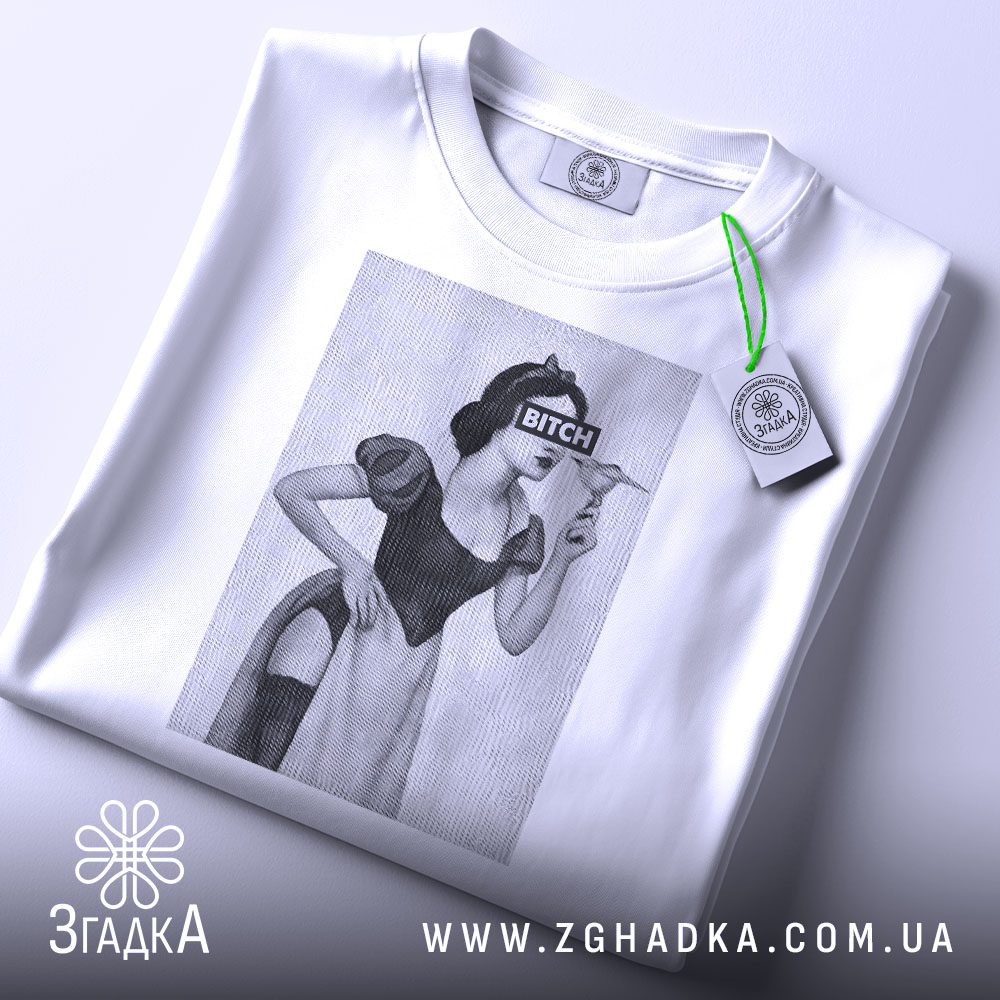 Футболка чорний гумор, біла, зображена зверху, на фоні світло-сірого кольору, принт з жінкою в костюмі, з написом 'BITCH', на футболці є етикетка з зеленим шнурком.