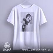 Футболка чорний гумор, біла, підвішена на вішалці, на фоні світло-сірого кольору, принт з жінкою в костюмі, з написом 'BITCH', видно деталі рукавів.