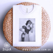 Футболка чорний гумор, біла, зображена зверху на плетеній кошику, на фоні світло-сірого кольору, принт з жінкою в костюмі, з написом 'BITCH', видно деталі горловини.