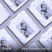 Футболка чорний гумор, біла, зображена з фронтального ракурсу, на фоні світло-сірого кольору, принт зображає жінку в костюмі, з написом 'BITCH' на обличчі.