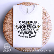 Футболка чудова донечка, біла, зображена зверху на плетеній кошику, фон світло-сірий, принт чорного кольору з жовтими зірками, текст українською мовою.