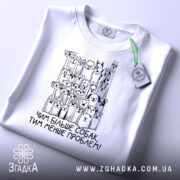 Футболка чим більше собак, біла, складена на поверхні, фон світлий, принт з малюнками собак і написом 'Чим більше собак, тим менше проблем!' у чорному кольорі, з зеленим ярликом.