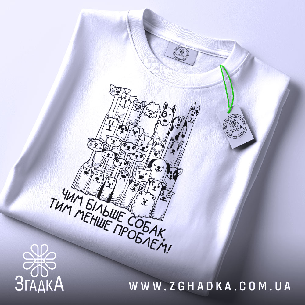 Футболка чим більше собак, біла, складена на поверхні, фон світлий, принт з малюнками собак і написом 'Чим більше собак, тим менше проблем!' у чорному кольорі, з зеленим ярликом.