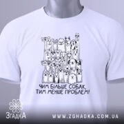 Футболка чим більше собак, біла, складена на поверхні, фон світлий, принт з малюнками собак і написом 'Чим більше собак, тим менше проблем!' у чорному кольорі, з зеленим ярликом.