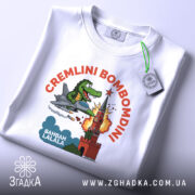 Футболка Cremlini Bombomdini білого кольору, складена на столі, ракурс зверху, фон світло-сірий, принт з крокодилом на літаку, що атакує червону вежу, з прикріпленою етикеткою.