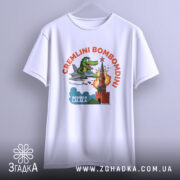 Футболка Cremlini Bombomdini білого кольору, підвішена на вішалці, ракурс спереду, фон світло-сірий, принт з крокодилом на літаку, що атакує червону вежу з вибухом.