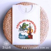 Футболка Cremlini Bombomdini білого кольору, складена на плетеній кошику, ракурс зверху, фон натуральний, принт з крокодилом на літаку, що атакує червону вежу, з яскравими деталями вибуху.