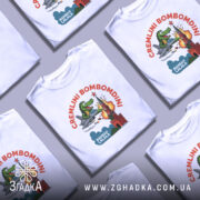 Футболка Cremlini Bombomdini білого кольору, зображена на манекені, ракурс спереду, фон світло-сірий, принт містить яскраву ілюстрацію крокодила на літаку, що атакує червону вежу з вибухом.