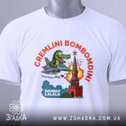 Футболка Cremlini Bombomdini білого кольору, складена на столі, ракурс зверху, фон світло-сірий, принт з крокодилом на літаку, що атакує червону вежу, з прикріпленою етикеткою.