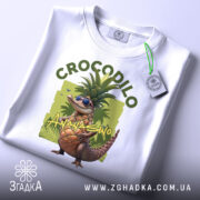 Футболка Crocodillo Ananasinno білого кольору, складена на поверхні, з видимим етикеткою, фон світло-сірий, принт з ящіркою в окулярах та ананасом, оточений зеленими елементами, розташована на рівній поверхні.