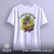 Футболка Crocodillo Ananasinno білого кольору, підвішена на вішалці, фон світло-сірий, принт з ящіркою в окулярах та ананасом, оточений зеленими елементами, зображена з фронтального ракурсу.