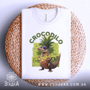 Футболка Crocodillo Ananasinno білого кольору, складена на плетеній основі, фон світлий, принт з ящіркою в окулярах та ананасом, оточений зеленими елементами, розташована на круглому плетеному підносі.