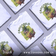 Футболка Crocodillo Ananasinno білого кольору, зображена з фронтального ракурсу на манекені, фон світло-сірий, принт з веселим ящіркою в окулярах та ананасом на голові, оточений зеленими елементами.