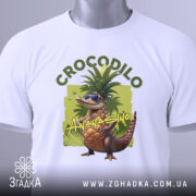 Футболка Crocodillo Ananasinno білого кольору, складена на поверхні, з видимим етикеткою, фон світло-сірий, принт з ящіркою в окулярах та ананасом, оточений зеленими елементами, розташована на рівній поверхні.
