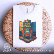 Футболка дайте спокій, біла, лежить на плетеній основі, фон світлий, принт з лягушкою на кріслі, текст 'Дайте мені спокій'.