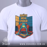 Футболка дайте спокій, біла, лежить на столі, фон світлий, принт з лягушкою на кріслі, з текстом 'Дайте мені спокій', з етикеткою на шнурку.