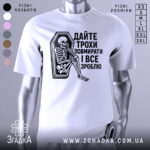 Футболка дайте трохи повмирати, біла, зображена з фронтального ракурсу, на манекені, фон світло-сірий, принт чорного кольору з малюнком скелета, що сидить у труні, текст українською мовою.