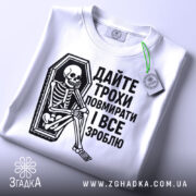 Футболка дайте трохи повмирати, біла, зображена з верхнього ракурсу, складена на поверхні, фон світло-сірий, принт чорного кольору з малюнком скелета у труні, текст українською мовою, на футболці є етикетка з зеленим шнурком.