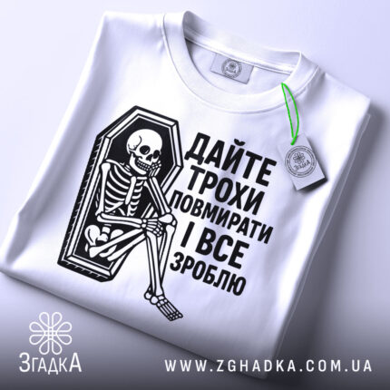 Футболка дайте трохи повмирати, біла, зображена з верхнього ракурсу, складена на поверхні, фон світло-сірий, принт чорного кольору з малюнком скелета у труні, текст українською мовою, на футболці є етикетка з зеленим шнурком.