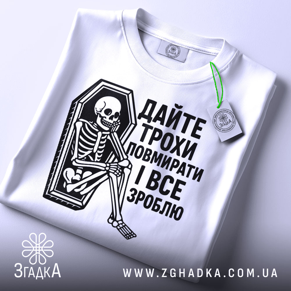 Футболка дайте трохи повмирати, біла, зображена з верхнього ракурсу, складена на поверхні, фон світло-сірий, принт чорного кольору з малюнком скелета у труні, текст українською мовою, на футболці є етикетка з зеленим шнурком.