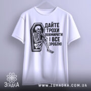 Футболка дайте трохи повмирати, біла, зображена з фронтального ракурсу, підвішена на вішалці, фон світло-сірий, принт чорного кольору з малюнком скелета, що сидить у труні, текст українською мовою.