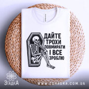 Футболка дайте трохи повмирати, біла, зображена з верхнього ракурсу, складена на плетеній основі, фон світлий, принт чорного кольору з малюнком скелета у труні, текст українською мовою.