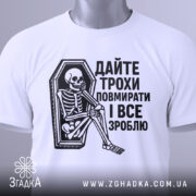 Футболка дайте трохи повмирати, біла, зображена з верхнього ракурсу, складена на поверхні, фон світло-сірий, принт чорного кольору з малюнком скелета у труні, текст українською мовою, на футболці є етикетка з зеленим шнурком.