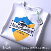 Футболка далекобійники України, білого кольору, складена на плоскій поверхні, фон світло-сірий, принт з синім та жовтим щитом, малюнком вантажівки та написом 'ДАЛЕКОБІЙНИКИ УКРАЇНИ', з зеленим ярликом.