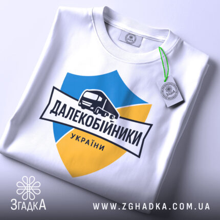 Футболка далекобійники України, білого кольору, складена на плоскій поверхні, фон світло-сірий, принт з синім та жовтим щитом, малюнком вантажівки та написом 'ДАЛЕКОБІЙНИКИ УКРАЇНИ', з зеленим ярликом.