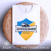 Футболка далекобійники України, білого кольору, складена на плоскій поверхні з плетеного кошика, фон білий, принт з синім та жовтим щитом, малюнком вантажівки та написом 'ДАЛЕКОБІЙНИКИ УКРАЇНИ'.