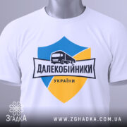 Футболка далекобійники України, білого кольору, складена на плоскій поверхні, фон світло-сірий, принт з синім та жовтим щитом, малюнком вантажівки та написом 'ДАЛЕКОБІЙНИКИ УКРАЇНИ', з зеленим ярликом.