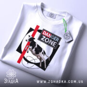 Футболка Danger Zone білого кольору, складена на поверхні, ракурс зверху, фон світлий, принт з коровою, червоною лінією та написом 'DANGER ZONE', з етикеткою на комірці.