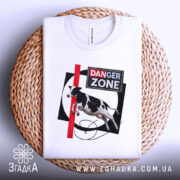 Футболка Danger Zone білого кольору, складена на плетеній основі, ракурс зверху, фон світлий, принт з коровою, червоною лінією та написом 'DANGER ZONE'.