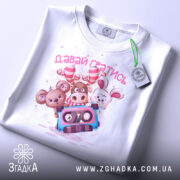 Футболка давай гратись, білого кольору, зображена зверху, складена на поверхні, фон світло-сірий, принт з чотирма мультяшними тваринами та касетою, текст 'давай гратись' виконаний рожевим кольором, на футболці є етикетка з зеленим шнурком.