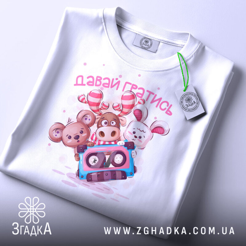 Футболка давай гратись, білого кольору, зображена зверху, складена на поверхні, фон світло-сірий, принт з чотирма мультяшними тваринами та касетою, текст 'давай гратись' виконаний рожевим кольором, на футболці є етикетка з зеленим шнурком.